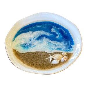 May include: A white oval dish featuring a resin art design that mimics a beach scene. The surface displays a sandy brown base with embedded seashells and a starfish, transitioning into swirling blue and white waves of ocean water.