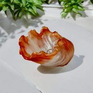 May include: A decorative, shell-shaped dish with a swirling pattern of orange and white. The dish has a glossy finish and appears to be made of resin or a similar material. A small, silver ring rests inside the dish.
