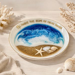 May include: A round white ceramic dish with a gold rim featuring a resin ocean scene with blue and white waves crashing onto a sandy beach. A small starfish and two seashells are embedded in the sand. The text reads: His favorite place became our forever tradition.