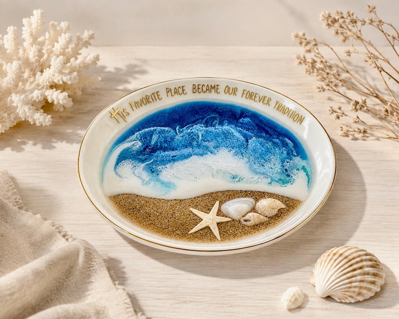 May include: A round white ceramic dish with a gold rim featuring a resin ocean scene with blue and white waves crashing onto a sandy beach. A small starfish and two seashells are embedded in the sand. The text reads: His favorite place became our forever tradition.