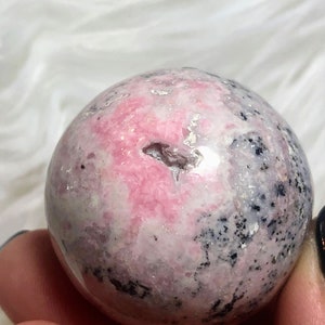 May include: A pink and grey sphere of polished rhodochrosite, a type of gemstone, with a natural crack in the surface.