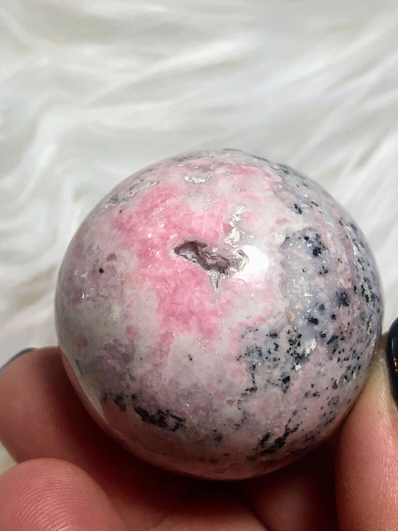 May include: A pink and grey sphere of polished rhodochrosite, a type of gemstone, with a natural crack in the surface.