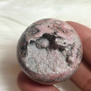 May include: A pink and black sphere-shaped crystal with a rough, textured surface. The crystal has a large opening revealing a white, crystalline interior.