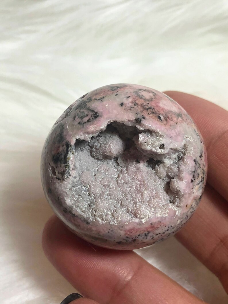 May include: A pink and black sphere-shaped crystal with a rough, textured surface. The crystal has a large opening revealing a white, crystalline interior.
