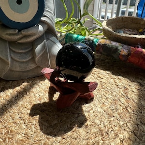 May include: A glossy black crystal ball sits on a small, reddish-brown wooden stand. The sphere reflects light, creating highlights. The stand has a three-legged design. The background includes a statue, plants, and a woven mat.