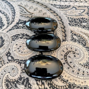 May include: Three glossy, black, oval-shaped decorative stones are arranged vertically. The stones reflect the surrounding environment, creating a mirror-like effect. The stones are displayed on a patterned fabric.
