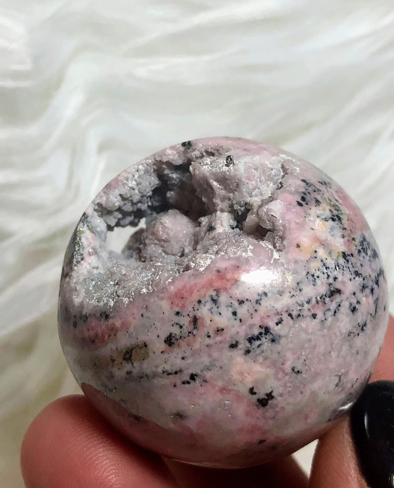 May include: A pink and black sphere with a crystal-like texture. The sphere has a large opening revealing a cavity with smaller crystals inside.