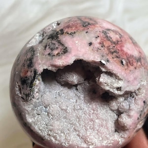 May include: A pink and black sphere of polished rhodochrosite with a natural druzy interior. The sphere has a rough, uneven surface and a large opening revealing the druzy crystals.