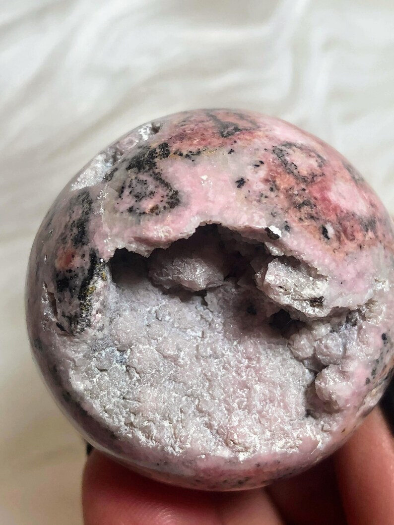 May include: A pink and black sphere of polished rhodochrosite with a natural druzy interior. The sphere has a rough, uneven surface and a large opening revealing the druzy crystals.