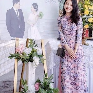 May include: A woman in a floral print dress stands near a wedding photo display and floral arrangements. The dress features a pink base with blue and purple floral patterns. A black purse hangs from her shoulder.