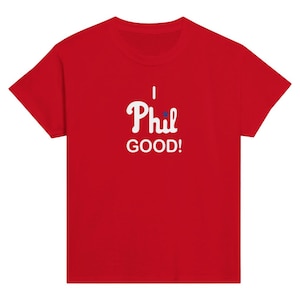 Op de afbeelding: Een felrood T-shirt met korte mouwen en witte tekst I Phil GOOD! op de borst. Een klein blauw sterretje vervangt de punt op de letter i in het woord Phil. Het kledingstuk is voorzien van een ronde halslijn.