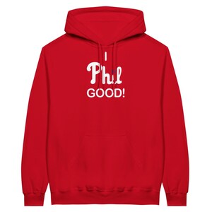 Op de afbeelding: Een felrode hoodie met een kangoeroezak aan de voorzijde en lange mouwen. Op de borst staat de witte tekst "I Phil GOOD!", waarbij de "i" in "Phil" is vervangen door een klein blauw grafisch element, gecentreerd op het kledingstuk.