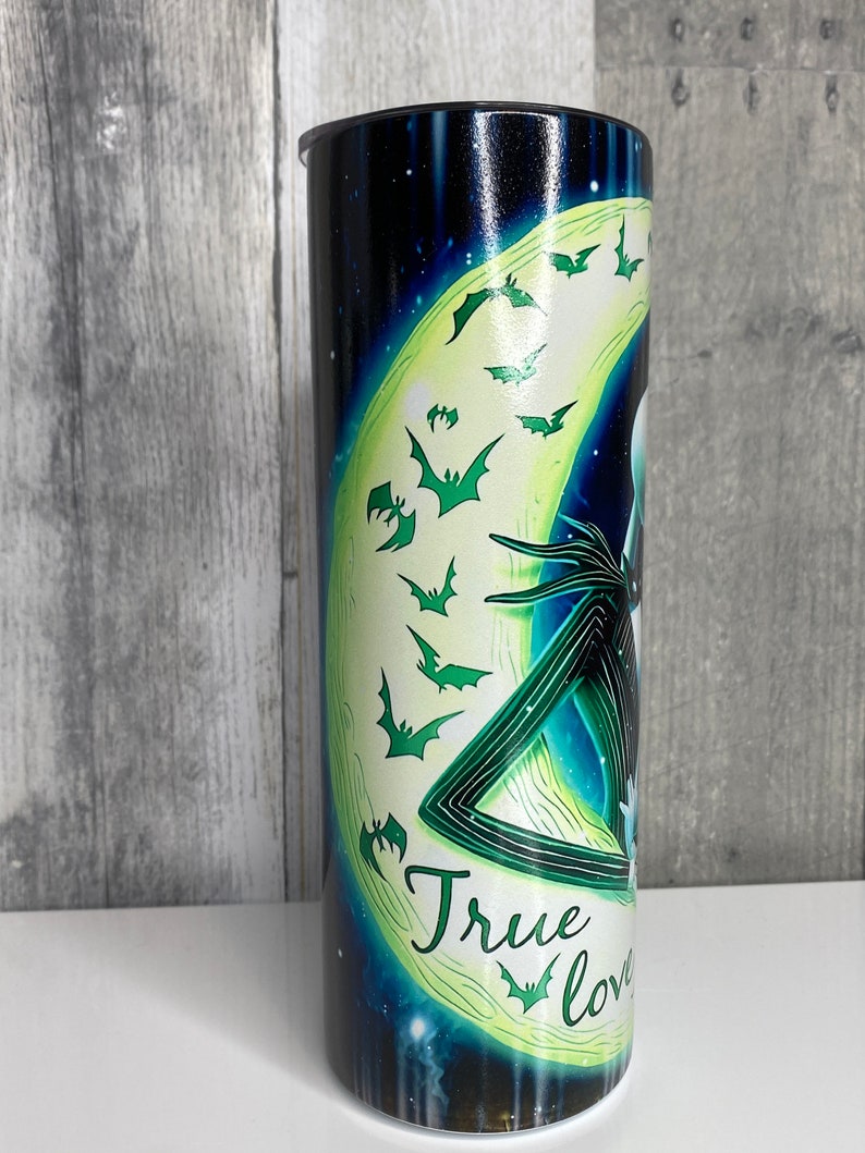 May include: A black and green stainless steel tumbler with a graphic of Jack Skellington and Sally from the Nightmare Before Christmas movie. The graphic features a crescent moon with bats flying around it. The text "True Love" is written on the tumbler.