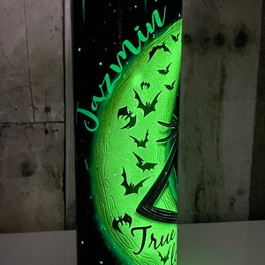 May include: A black and green glow-in-the-dark tumbler with a moon and bats design. The name "Jazmin" is written on the moon. The text "True" is written below the moon.