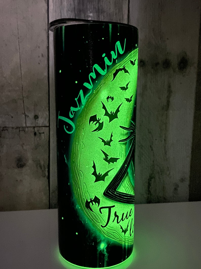 May include: A black and green glow-in-the-dark tumbler with a moon and bats design. The name "Jazmin" is written on the moon. The text "True" is written below the moon.