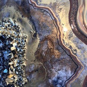 Op de afbeelding: Abstracte harskunst in een gouden, bruine en zwarte kleurstelling. Het hars heeft een textuur, gelaagde look met een glinsterende afwerking. De kunst toont een cluster van zwarte en gouden stenen in de linkerbenedenhoek.