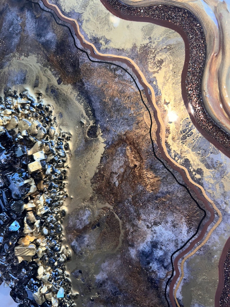 Op de afbeelding: Abstracte harskunst in een gouden, bruine en zwarte kleurstelling. Het hars heeft een textuur, gelaagde look met een glinsterende afwerking. De kunst toont een cluster van zwarte en gouden stenen in de linkerbenedenhoek.
