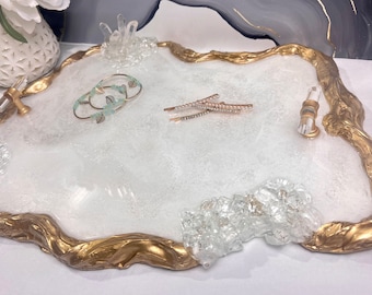May include: A decorative tray with a white and gold design. The tray has a gold-colored, organic-shaped border and is adorned with clear crystal accents. Two hair clips and a bracelet are on the tray.