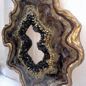 Op de afbeelding: Een groot, abstract kunstwerk van hars in de vorm van een agaatgeode. De geode is geschilderd in tinten bruin, goud en zwart, met een grote opening in het midden. De geode is omgeven door een gouden lijst.