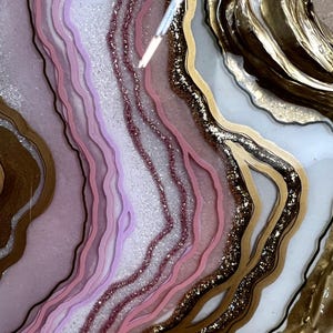 K&ouml;nnte beinhalten: Abstraktes Kunstwerk mit geschichtetem Kunstharz in Rosa, Lavendel und Wei&szlig;, akzentuiert mit Gold und Glitzer. Das Design &auml;hnelt einer Geode oder einem Achatschnitt mit flie&szlig;enden, organischen Formen.