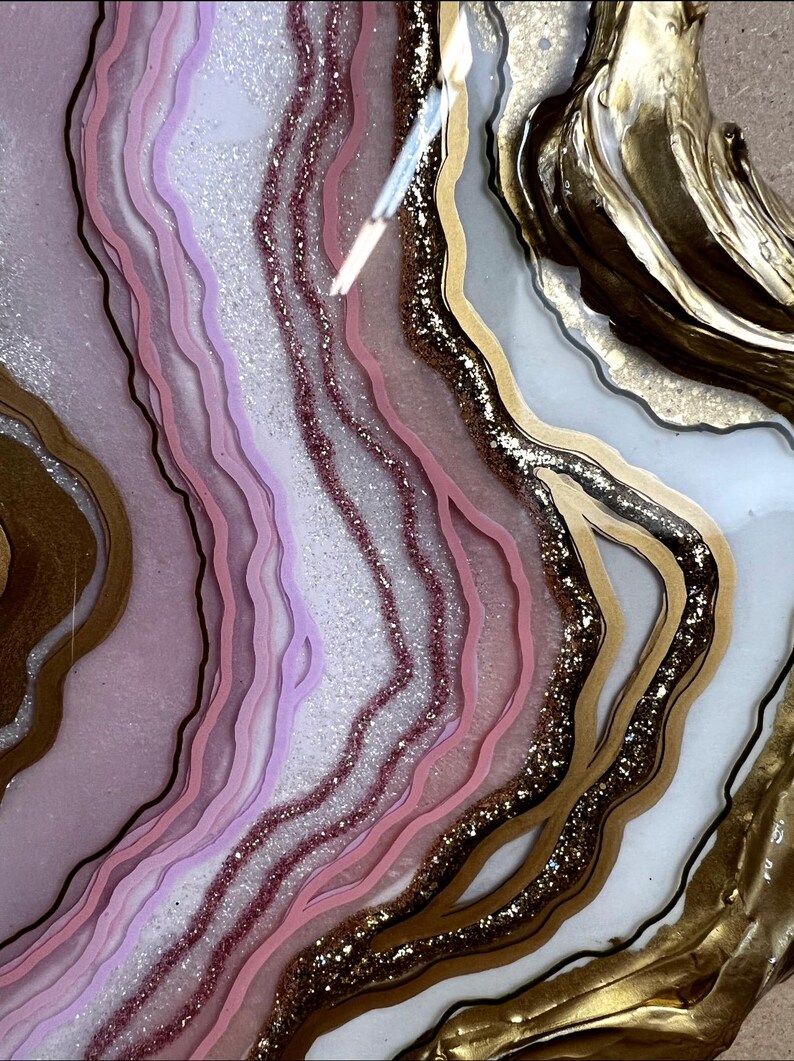 K&ouml;nnte beinhalten: Abstraktes Kunstwerk mit geschichtetem Kunstharz in Rosa, Lavendel und Wei&szlig;, akzentuiert mit Gold und Glitzer. Das Design &auml;hnelt einer Geode oder einem Achatschnitt mit flie&szlig;enden, organischen Formen.