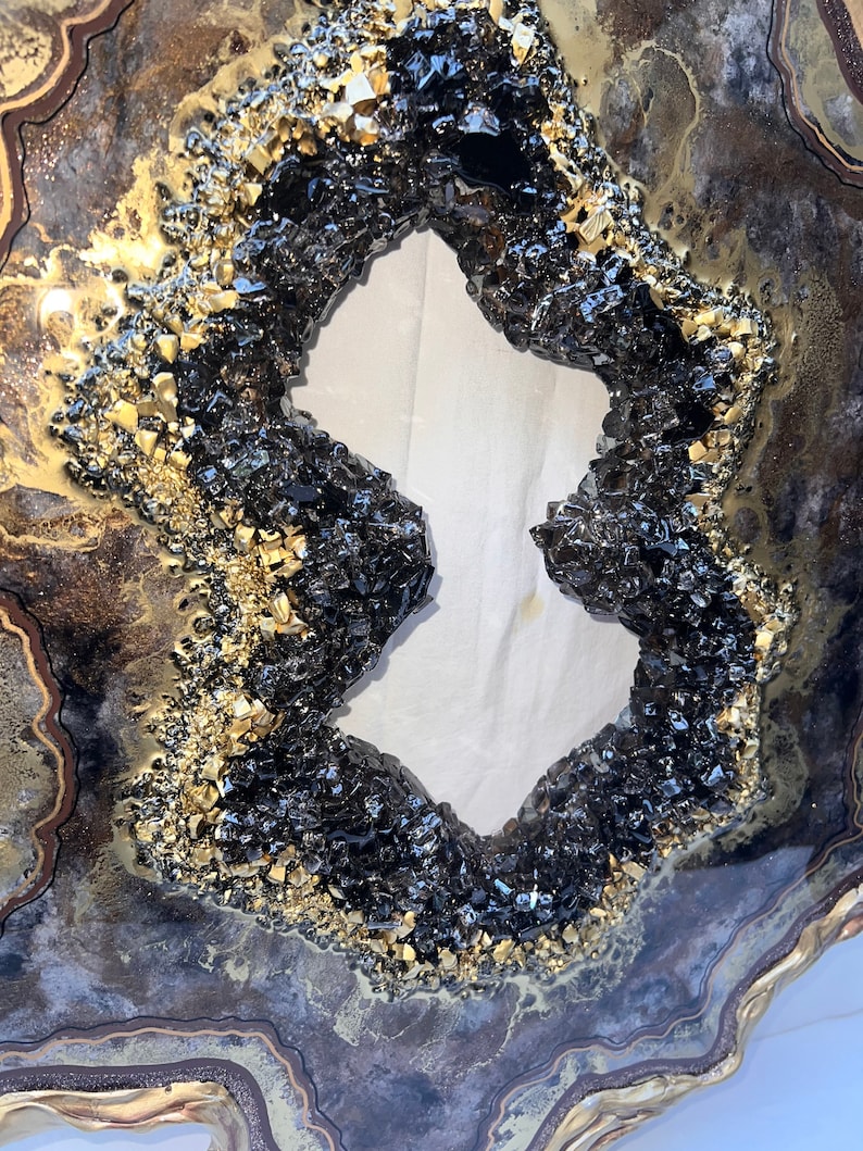 Op de afbeelding: Een groot, rond, kunsthars kunstwerk met een zwart en gouden agaatontwerp. De agaat heeft een groot, open midden, waardoor een witte achtergrond zichtbaar wordt.