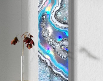 Puede incluir: Una pintura abstracta vertical con remolinos y purpurina azules, blancos y plateados. La pintura está sobre lienzo y está colgada en una pared blanca.