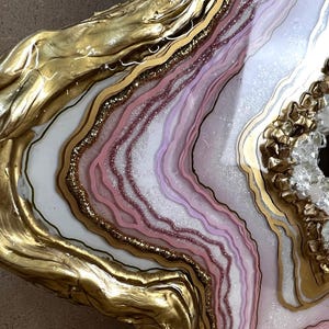 K&ouml;nnte beinhalten: Nahaufnahme einer dekorativen Geodenscheibe mit goldfarbenen R&auml;ndern. Das Innere zeigt Schichten aus wei&szlig;en, rosa und lila Farbt&ouml;nen mit Glitzerakzenten. Ein kristallgef&uuml;llter Hohlraum sorgt f&uuml;r Textur und visuelles Interesse.