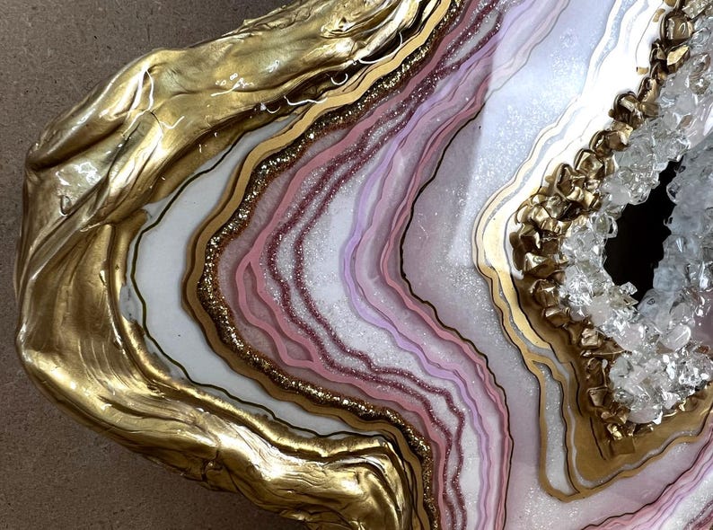 K&ouml;nnte beinhalten: Nahaufnahme einer dekorativen Geodenscheibe mit goldfarbenen R&auml;ndern. Das Innere zeigt Schichten aus wei&szlig;en, rosa und lila Farbt&ouml;nen mit Glitzerakzenten. Ein kristallgef&uuml;llter Hohlraum sorgt f&uuml;r Textur und visuelles Interesse.