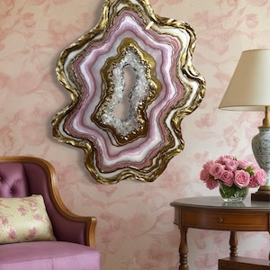 K&ouml;nnte beinhalten: Ein gro&szlig;er, dekorativer Wandspiegel in Form einer Geode. Der Spiegel hat Schichten aus Wei&szlig;, Rosa und Gold mit einem kristallartigen Zentrum. Der Rahmen hat einen unregelm&auml;&szlig;igen Goldrand. Der Spiegel ist auf einer rosa Blumentapete angebracht.