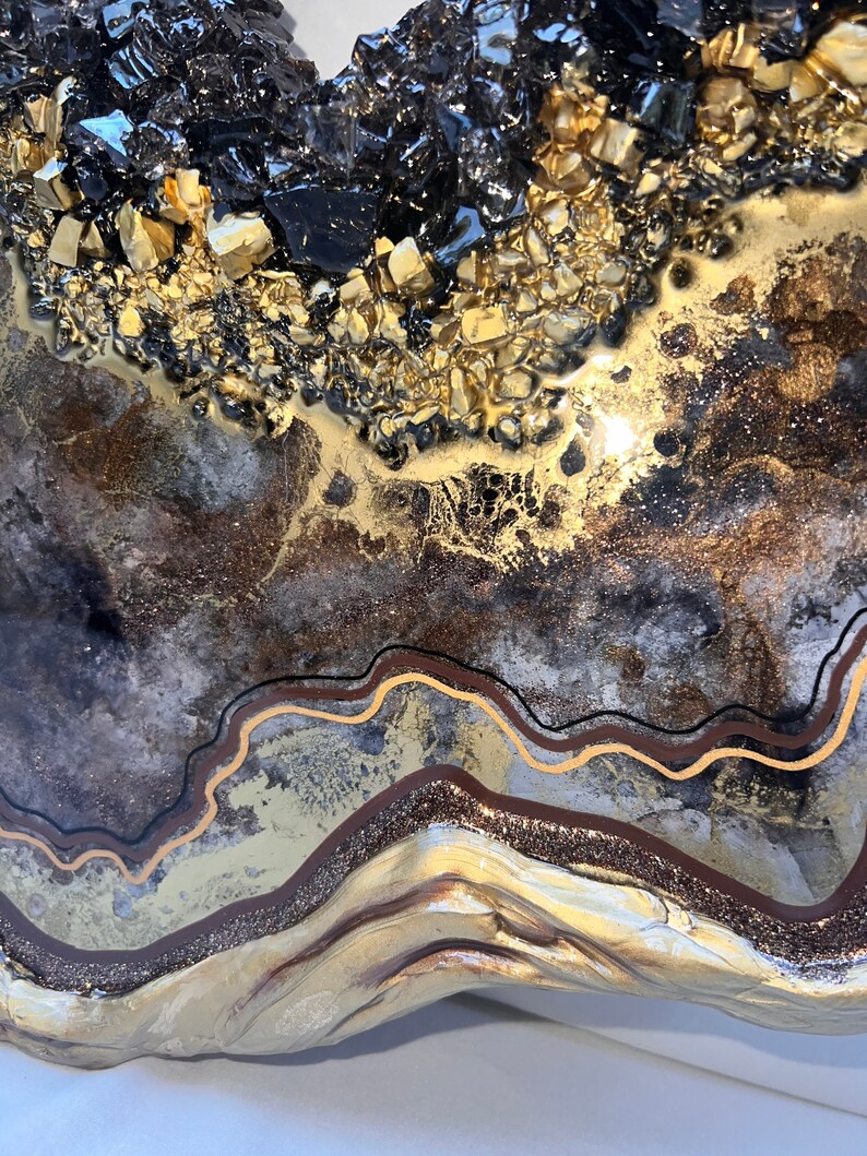 Op de afbeelding: Abstracte harskunst met een gouden en zwarte kleurstelling. Het hars is gelaagd en getextureerd, met een gouden glitterafwerking. De kunst is in de vorm van een hart.