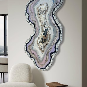 May include: Large, abstract wall art piece resembling a geode, featuring concentric layers of color. The artwork has a wavy, irregular shape with shades of lavender, gray, white, and gold. The outer edge is textured with a dark, speckled border.