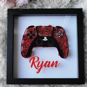 May include: Framed artwork featuring a black and red PlayStation controller design. The controller is filled with red rose details and the name "Ryan" in red script. The artwork is set against a white background within a black frame.