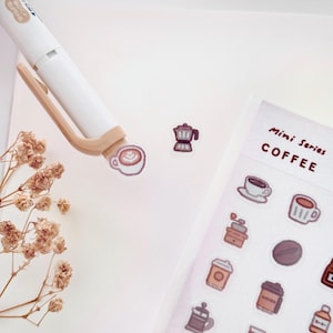 Può includere: Un foglio di adesivi a tema caffè con varie illustrazioni, tra cui tazze di caffè, un macinacaffè e una caffettiera. Le parole "Mini Series COFFEE" sono stampate sul foglio. Una penna bianca e fiori secchi sono anche nell'immagine.