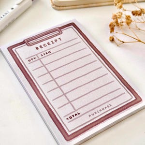 May include: A rectangular notepad with a receipt design. The notepad features a light pink border and the word "RECEIPT" at the top. The notepad has lines for quantity and item descriptions. The word "TOTAL" is at the bottom, with "PURINNHARI" next to it.