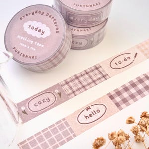 Könnte beinhalten: Masking Tape Rollen und Streifen in hellbraunen Farbtönen. Die Rollen haben ein Karomuster und die Aufschrift "today" und "everyday patterns". Die Streifen zeigen die Wörter "to-do", "cozy" und "hello".