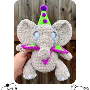 May include: A crocheted elephant toy with a jester theme. The elephant is light gray with large ears, blue-rimmed eyes, and a trunk. It wears a green and purple jester hat and a purple and green collar. The text "THE ELEPHANT JESTER" is at the top.