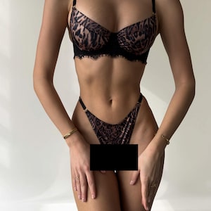 May include: A two-piece lingerie set featuring a leopard print design. The bra has black lace trim and adjustable straps. The matching thong completes the set. The set is designed with a brown and black animal print.