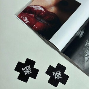 May include: Two black cross-shaped nipple covers with the text "FUCK ME!" in white. A magazine is open in the background, featuring a close-up of glossy red lips. The image is shot from above.