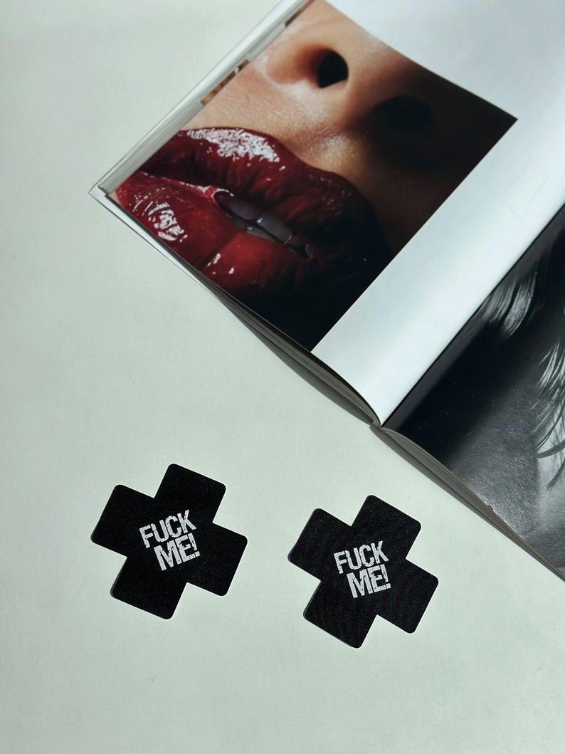 May include: Two black cross-shaped nipple covers with the text "FUCK ME!" in white. A magazine is open in the background, featuring a close-up of glossy red lips. The image is shot from above.