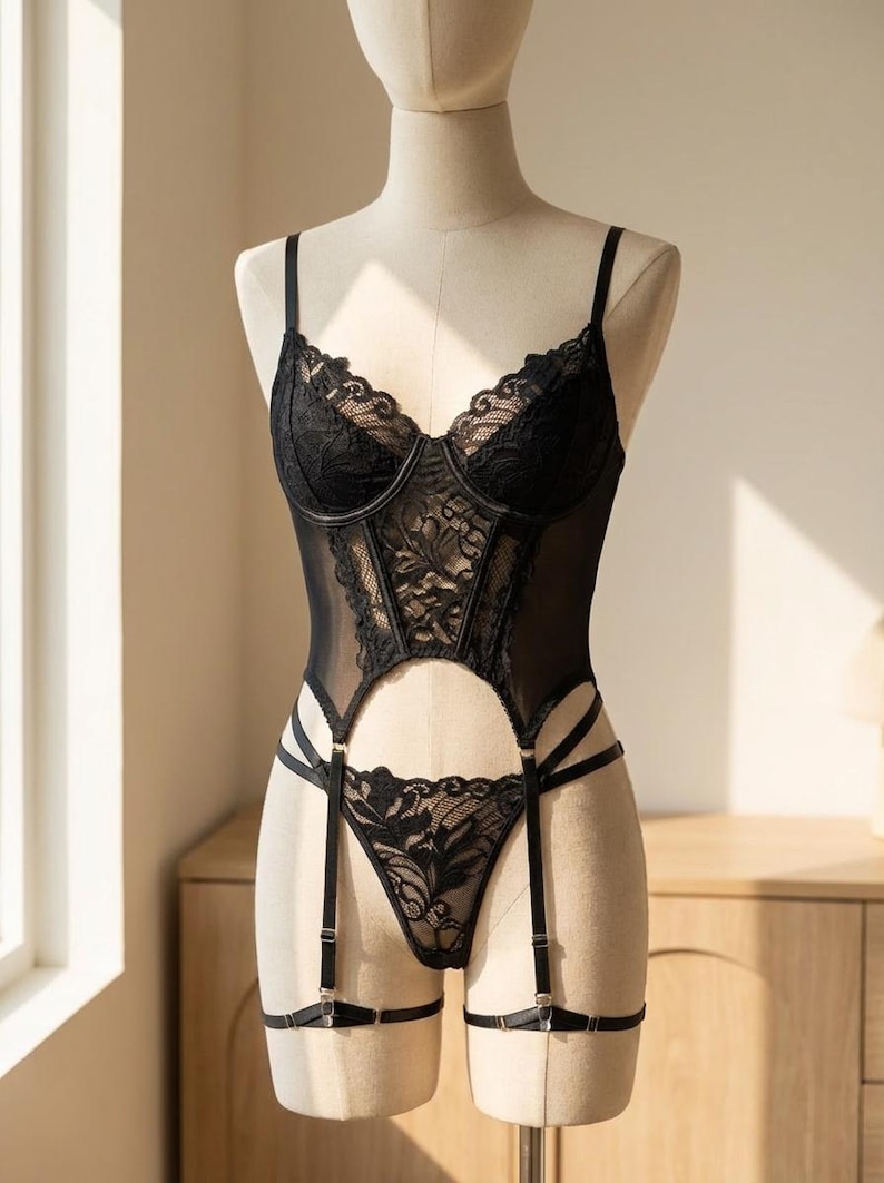 Puede incluir: Conjunto de lencer&iacute;a negra sobre un maniqu&iacute;. El conjunto incluye un bustier con detalles de encaje, tirantes ajustables y un tanga a juego con ligas. El bustier tiene un escote coraz&oacute;n y detalles de encaje floral.
