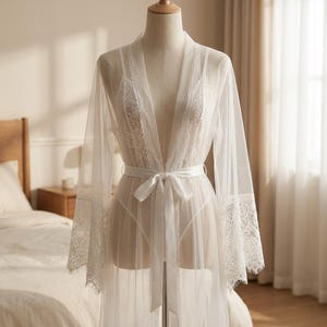 May include: A sheer white robe with lace detailing on the sleeves and bodice, tied at the waist with a matching satin ribbon. The robe is displayed on a mannequin, showcasing its delicate, flowing design. The garment is part of a lingerie collection.
