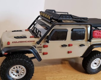 May include: Tan and black toy Jeep with the word "RUBICON" on the hood. The toy has black tires, a black roof rack with lights, and a red container on the back. The Jeep is a detailed model.