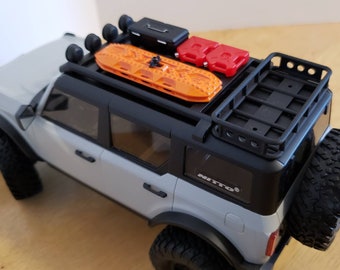 May include: A white Ford Bronco with a black roof rack, two red fuel cans, and an orange traction board. The vehicle has black wheels and tires. The roof rack has a black metal basket and a black toolbox.