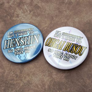May include: Two round buttons with text. One is blue with "HENSHIN" and the text "I'M NONBINARY BUT I HAVE TO" and "SO IDRC ABOUT THAT RN". The other is white with "OHGAI BUSOU" and the text "I'M NONBINARY BUT I HAVE TO" and "SO IDRC ABOUT THAT RN".