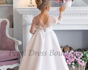 May include: A white satin dress with a sheer lace bodice and long sleeves. The dress has a full skirt and a button closure in the back. The dress is decorated with delicate floral lace appliques.