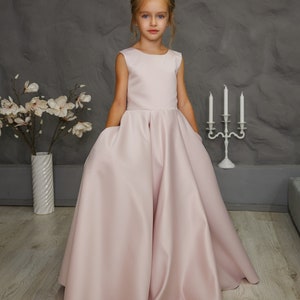 Pu&ograve; includere: Una bambina indossa un abito senza maniche in raso rosa chiaro con gonna ampia. L'abito ha tasche sui lati.