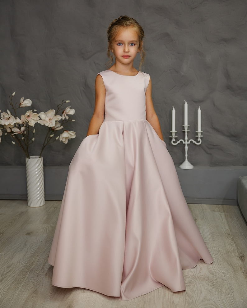 Pu&ograve; includere: Una bambina indossa un abito senza maniche in raso rosa chiaro con gonna ampia. L'abito ha tasche sui lati.