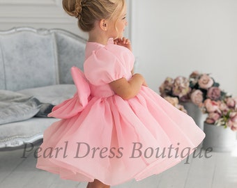 May include: A young girl wearing a pink tulle dress with a large bow in the back. The dress has a puffed sleeve and a fitted bodice. The girl is standing on a light wood floor. "Pearl Dress Boutique" is written in white text on the floor.