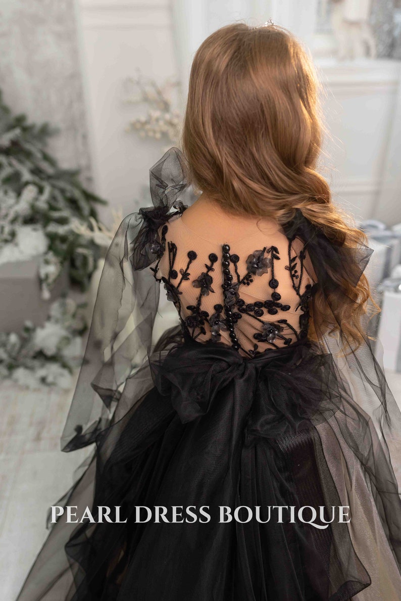 May include: A black tulle dress with a sheer lace bodice and a large bow at the back. The dress has a floral design and is adorned with pearl-like embellishments. The text "PEARL DRESS BOUTIQUE" is visible at the bottom of the image.